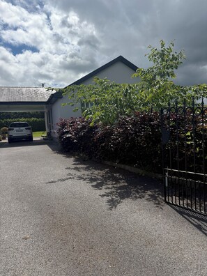 Uma entrada residencial com um carro estacionado, uma casa branca e uma árvore com folhas verdes e vermelhas.