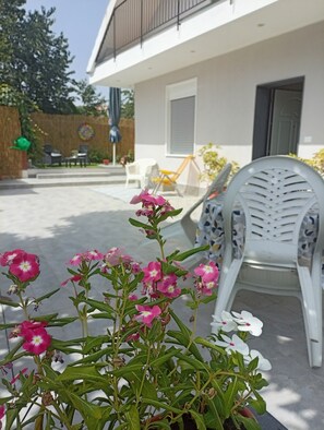 Un espacio al aire libre con muebles de exterior, una planta en maceta con flores rosas y un edificio blanco al fondo.