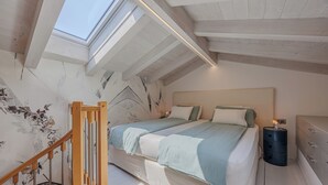 Un dormitorio con tragaluz, una cama con muchos almohadones, una mesita de noche y una mesita auxiliar.