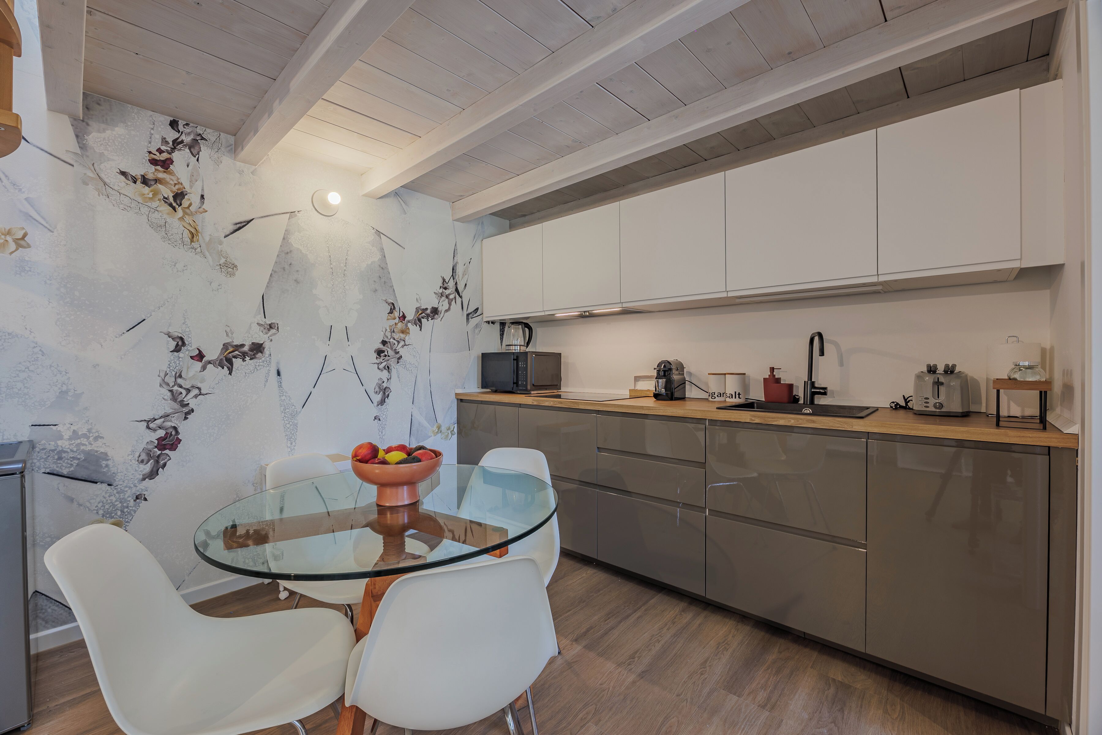 A modern kitchen with a glass dining table, white chairs, and a fruit bowl.