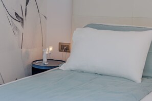 Ein Bett mit weißem Kopfkissen und blauer Decke, ein Nachttisch mit Lampe sowie eine Wand mit einem unvollständigen Gemälde.
