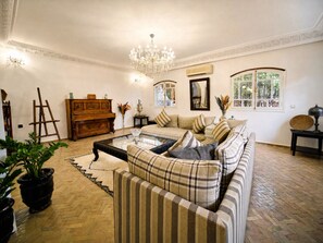 Ein Wohnzimmer mit einer Couch, einem Couchtisch, einer Chandelüster, einem Klavier und einem Gemälde an der Wand.