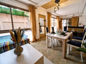 A dining area with a table set for two, a vase with dried plants, and a view of a swimming pool through large windows.