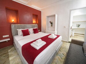 A hotel room with a large bed, red pillows, a white headboard, a red throw blanket, and a white bedside table with a vase.