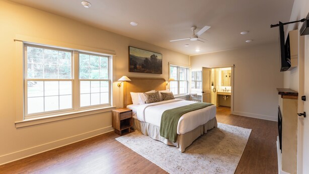 Une chambre avec un grand lit, une table de chevet, une lampe fixée au mur, un ventilateur de plafond et une fenêtre donnant sur des arbres.