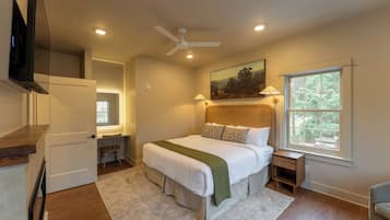 A bedroom with a bed, a desk, a chair, a TV, a ceiling fan, and a window with a view of trees.