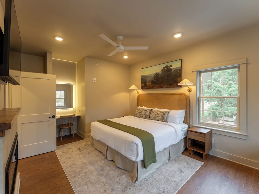 A bedroom with a bed, a desk, a chair, a TV, a ceiling fan, and a window with a view of trees.
