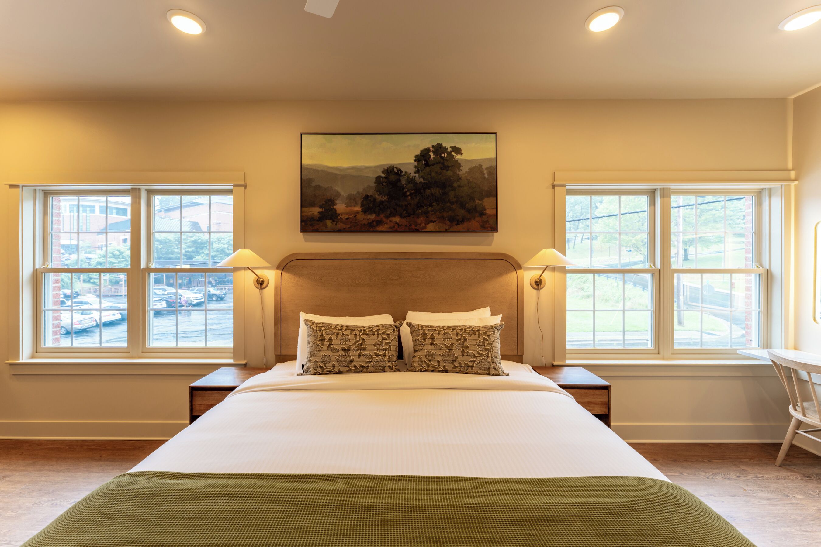 Une chambre à coucher avec un grand lit, deux tables de chevet, un tableau au-dessus du lit et des fenêtres de part et d’autre.