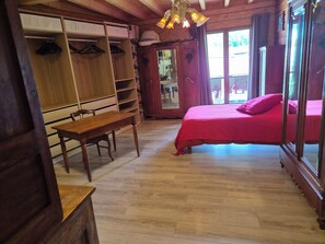 A room with a wooden floor, a bed with a red bedspread, a wooden desk with a chair, and a wardrobe with hanging space. - Gîte Le Rucan Atypical house in the Pyrenees Commingeoises (Montastruc-de-Salies)