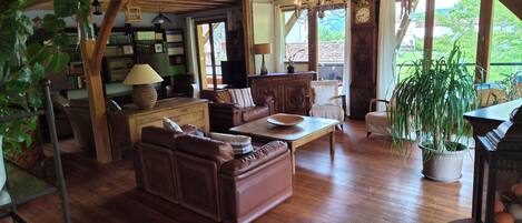 A cozy living room with wooden flooring, leather furniture, a piano, and a large window offering a view of the outdoors.