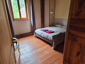 Un dormitorio con piso de madera, una cama con colchón gris y dos almohadas, una ventana con cortinas y una mesita de noche con lámpara.