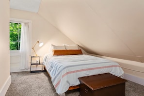 Un dormitorio con techo inclinado, una cama con ropa de cama a rayas, una mesita de noche con lámpara y una ventana con cortinas.