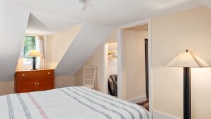 A bedroom with a sloped ceiling, a bed with striped bedding, a nightstand with a lamp, a wardrobe, and a walk-in closet.