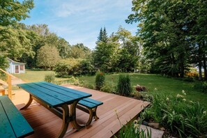 Una terraza de madera con una mesa de picnic y bancos, rodeada de abundante vegetación y árboles.