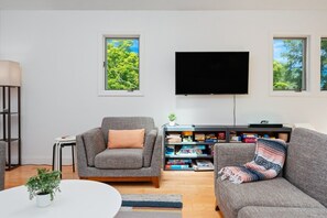 Un salón con un sofá gris, un sillón gris, un televisor de pantalla plana, una estantería con juegos de mesa y una ventana con vistas a árboles.