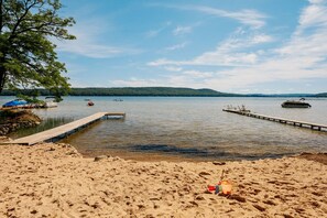 A sandy beach with a wooden dock extending into calm water, surrounded by trees and hills in the background.