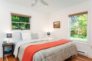 Quarto com cama, duas mesas de cabeceira, um ventilador de teto e duas janelas com vista para o verde.