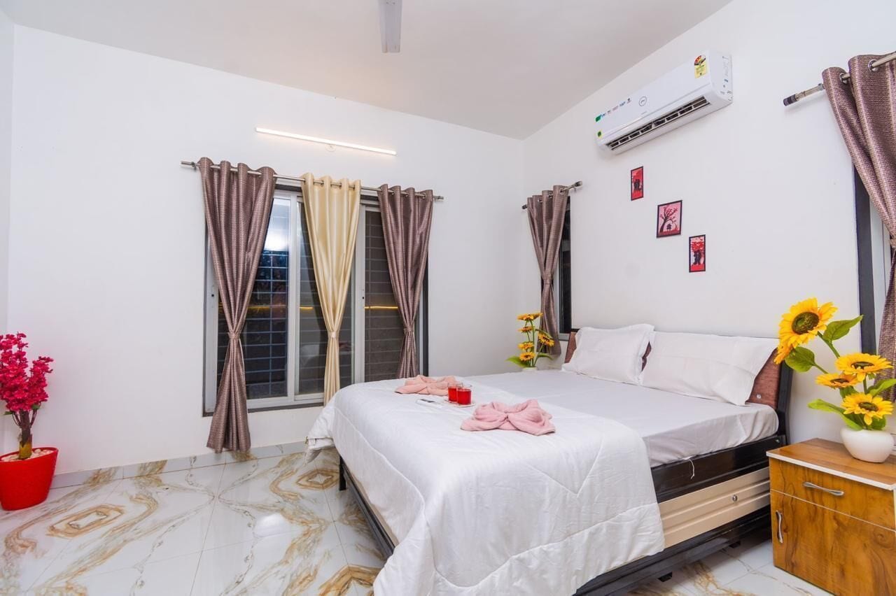 A hotel room with a bed, bedside table, air conditioning unit, curtains, a potted plant, and a vase with flowers.
