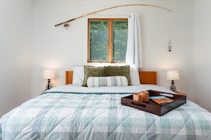 A neatly made bed with a checkered bedspread, two bedside lamps, a bedside table with a tray of books and mugs, and a window with a curtain.