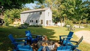 Un patio trasero con un brasero, sillas Adirondack azules y una casa al fondo.