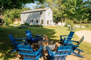 A backyard with a fire pit, blue Adirondack chairs, and a house in the background. - Camp Haven | Sandpiper Cottage | 4BR Beachfront | Glen Lake (Empire)