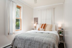 A bedroom with a bed, two bedside tables, a nightstand, a window with curtains, and a wall-mounted mirror. - Camp Haven | Sandpiper Cottage | 4BR Beachfront | Glen Lake (Empire)