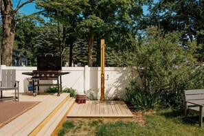 A backyard with a barbecue grill, a wooden deck, and a white fence. - Camp Haven | Sandpiper Cottage | 4BR Beachfront | Glen Lake (Empire)