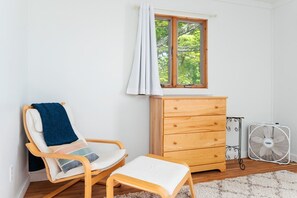 A room with a wooden rocking chair, a wooden dresser, a standing fan, and a window with curtains. - Camp Haven | Sandpiper Cottage | 4BR Beachfront | Glen Lake (Empire)