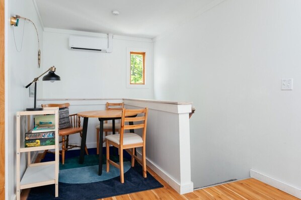 Habitación de esquina con una mesa y sillas de madera, una estantería, una lámpara de escritorio y una ventana con vistas a árboles.