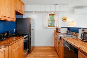 A compact kitchen with wooden cabinets, a black refrigerator, a stove, and a coffee maker. - Camp Haven | Sandpiper Cottage | 4BR Beachfront | Glen Lake (Empire)