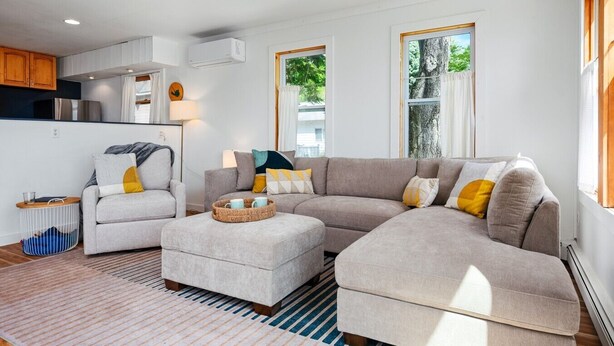Una sala moderna con un sofá modular, un sillón pequeño y una mesa de centro. Hay una zona de cocina con armarios y un equipo de aire acondicionado.
