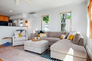 Ein modernes Wohnzimmer mit Ecksofa, einem Sessel und einem Couchtisch. Es gibt eine Küchenzeile mit Schränken und einer Klimaanlage.