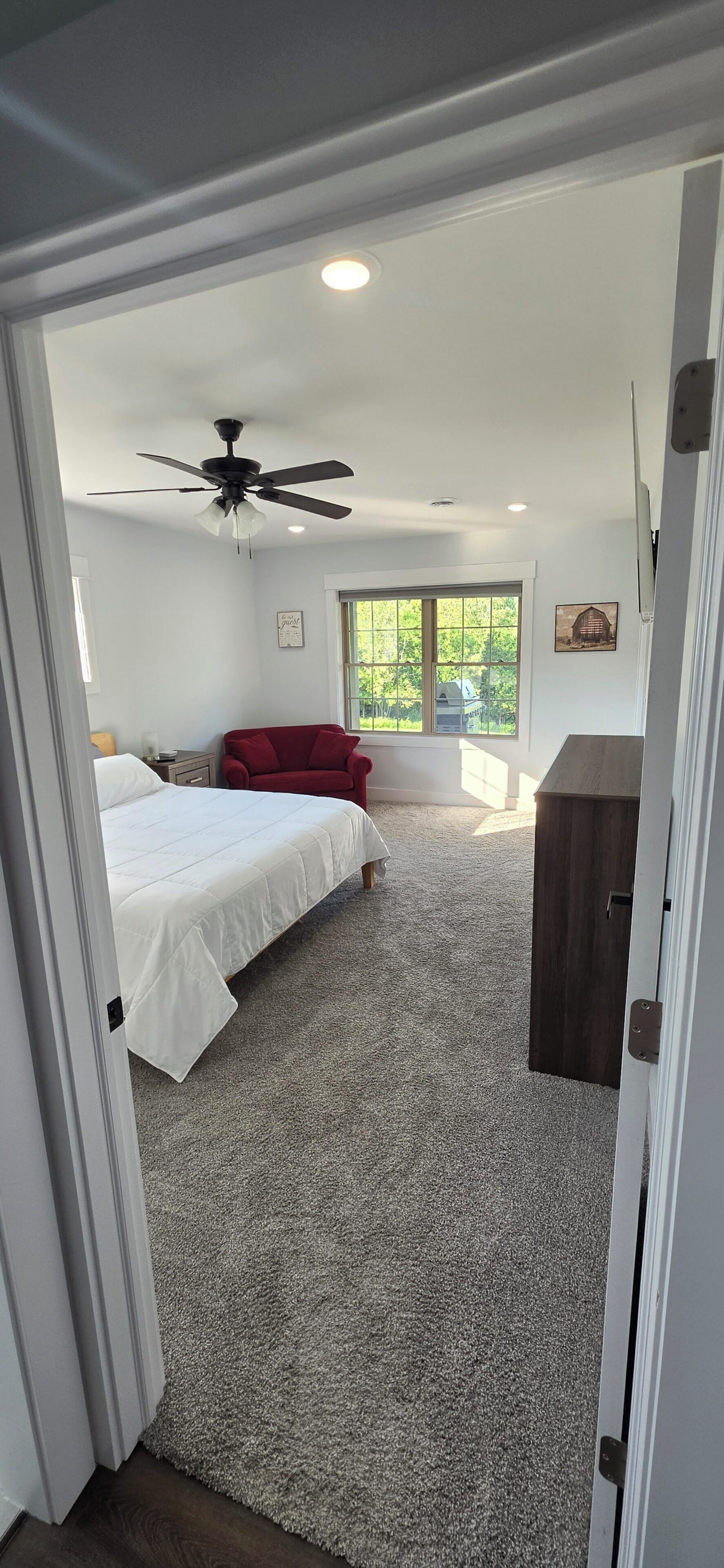 A bedroom with a bed, a red chair, a ceiling fan, and a window with a view of greenery.
