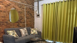 Ein Wohnzimmer mit grauem Sofa, gemusterten Kissen, einem Couchtisch, einem Teppich mit Muster, einem Holznachttisch und einem grünen Vorhang.