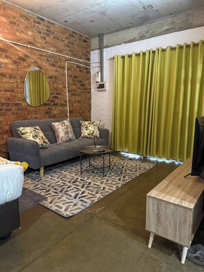 Ein Wohnzimmer mit grauem Sofa, gemusterten Kissen, einem Couchtisch, einem Teppich mit Muster, einem Holznachttisch und einem grünen Vorhang.
