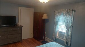 A room with a wooden dresser, a television, a door, a lamp, and a window with curtains. - Barr Street Retreat Charming 2BR Near Downtown Salem (Salem)