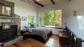 A room with a stone fireplace, a bed with green and brown bedding, a window with a view of trees, and a skylight. - Retromoderninthewoods steps from the best trails in the Linville gorge wildernes (Newland)