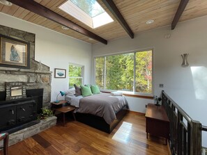 A room with a stone fireplace, a bed with green and brown bedding, a window with a view of trees, and a skylight. - Retromoderninthewoods steps from the best trails in the Linville gorge wildernes (Newland)
