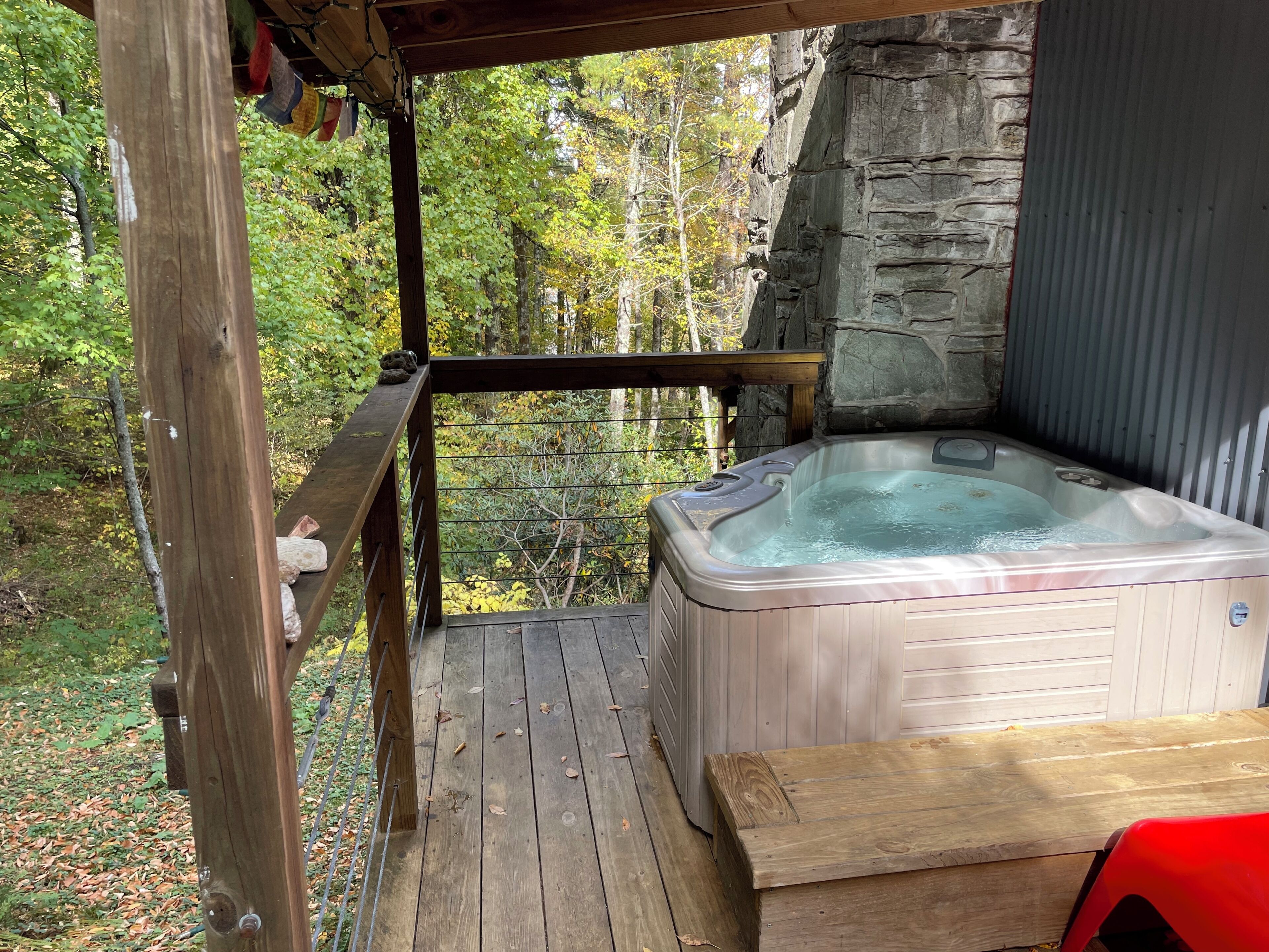A hot tub on a wooden deck with a stone wall backdrop and a view of a forest.