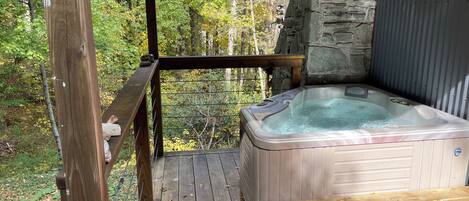 A hot tub on a wooden deck with a stone wall backdrop and a view of a forest.