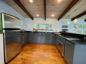 Uma cozinha moderna com frigorífico de aço inoxidável, soalho em padrão herringbone de madeira e uma janela com vista para o verde.