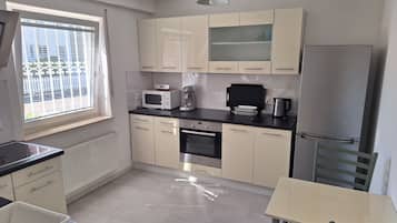Eine kompakte Küche mit Mikrowelle, Toaster, Wasserkocher und Backofen. Ein Fenster mit Jalousien, ein Heizkörper und ein Tisch mit Stühlen sind vorhanden.