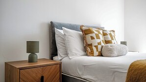 Ein ordentlich bezogenes Bett mit grauem Kopfteil, weißer Bettwäsche und gemusterten Kissen. Ein Nachttisch aus Holz mit Lampe und Schublade steht daneben.