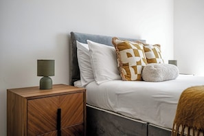 A neatly made bed with a grey headboard, white bedding, and patterned pillows. There is a wooden nightstand with a lamp and a drawer. - Canary Marina – Luxury 3‑Bed Apartment (Greater London)