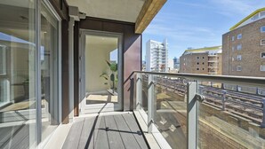 Ein Balkon mit Holzboden, Glastüren zu einem Raum und Blick auf eine Stadtlandschaft mit Gebäuden.