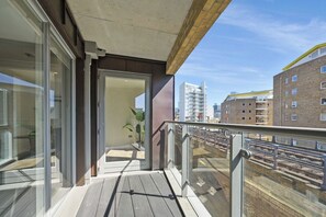 A balcony with a wooden floor, glass doors leading to a room, and a view of a cityscape with buildings. - Canary Marina – Luxury 3‑Bed Apartment (Greater London)