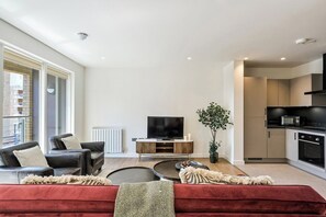 A modern living room with a red sofa, black leather chairs, a flat-screen TV, and a dining area with a microwave. - Canary Marina – Luxury 3‑Bed Apartment (Greater London)