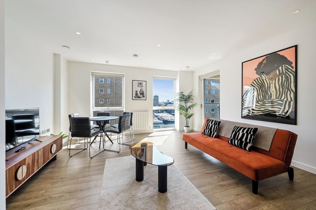 A modern living room with a sofa, coffee table, and TV stand. There is a dining area with chairs and a table. The room has large windows and a view of the city.