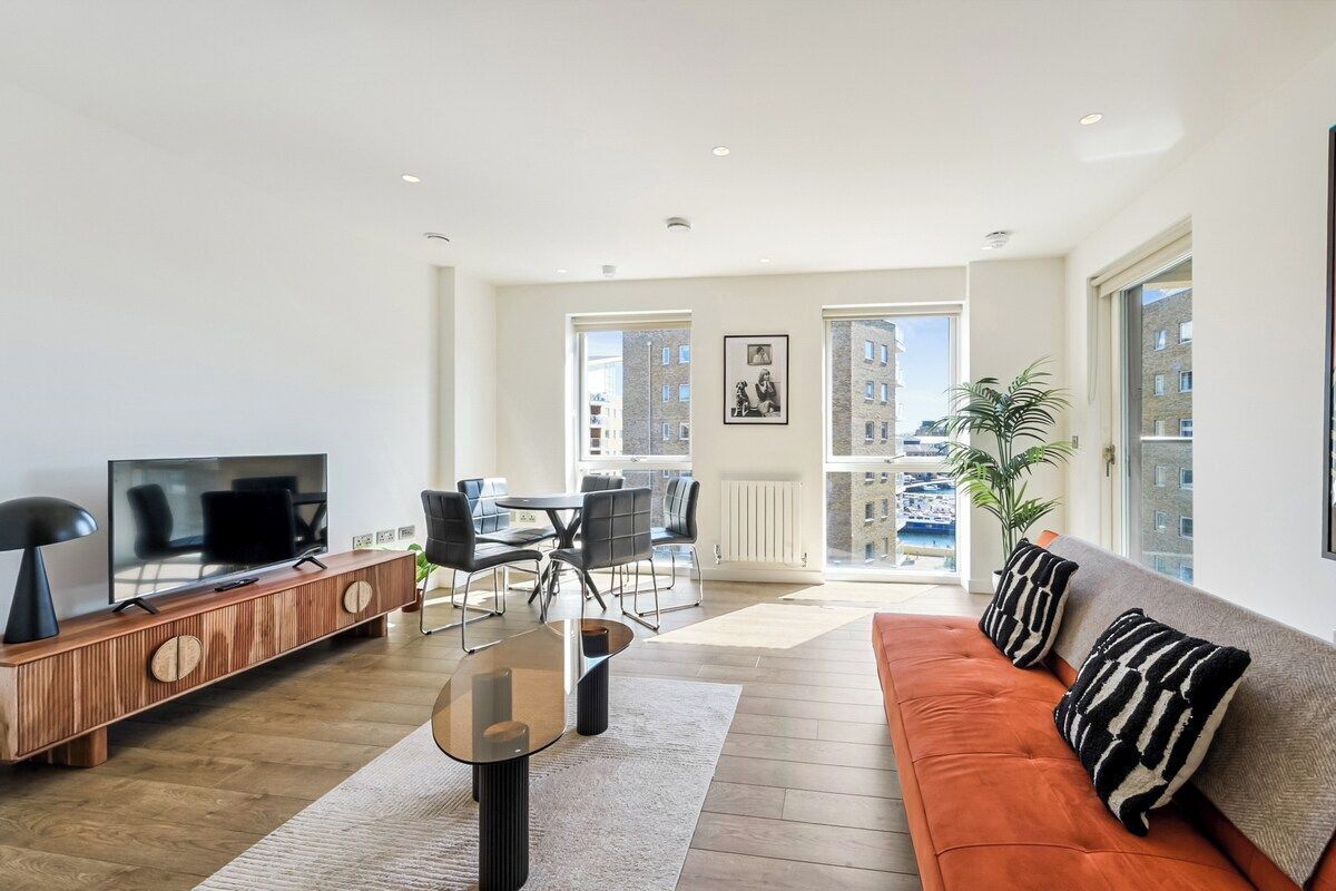 Een moderne woonkamer met een bank, televisie, eettafel, stoelen en een uitzicht op gebouwen door grote ramen.