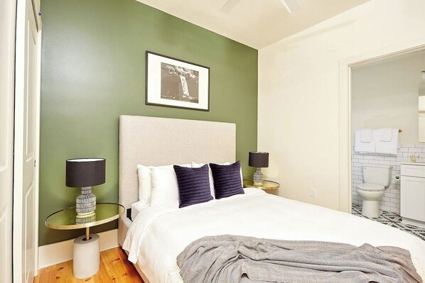 Ein Schlafzimmer mit Bett, zwei Nachttischen, einem Schminktisch und einem gerahmten Bild an der Wand. Ein Badezimmer ist durch eine offene Tür sichtbar.